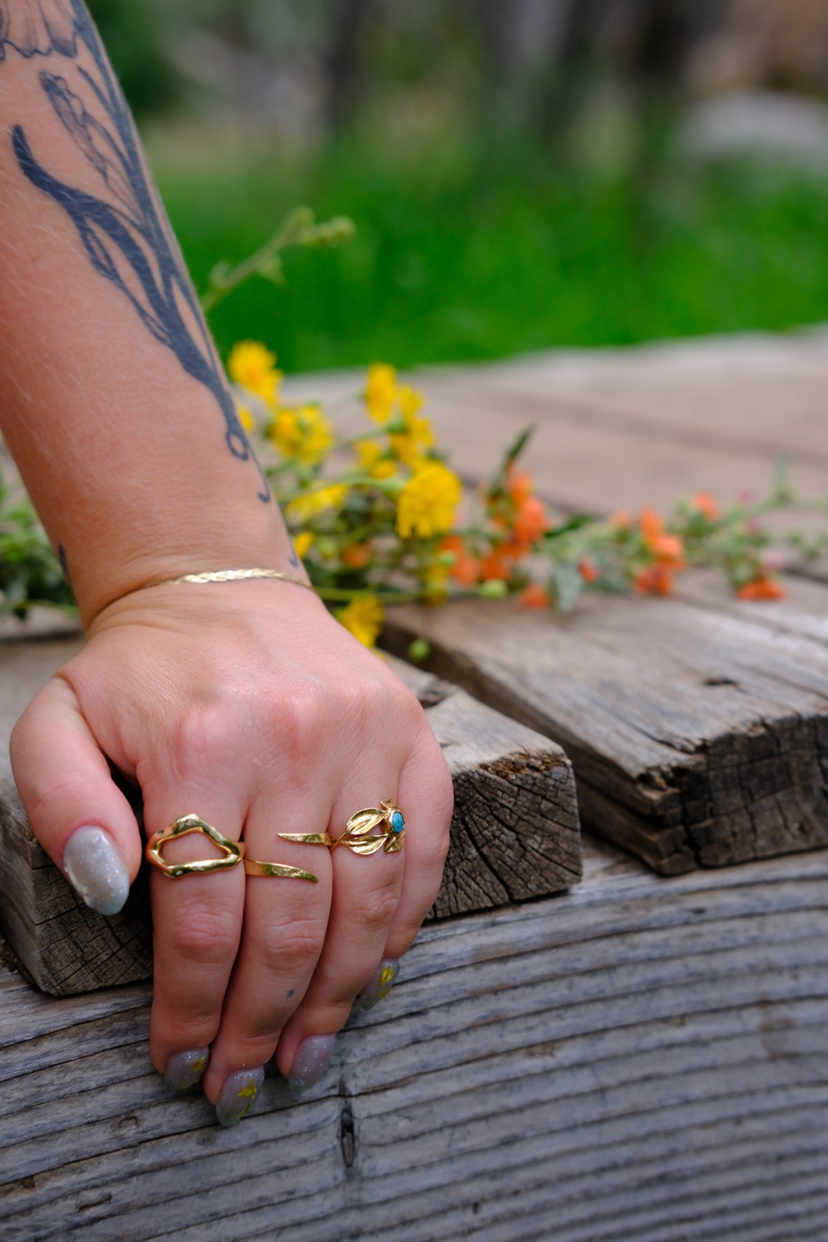 Model wears Snake Spiral Ring in 14k Gold, Split Root Ring in 14k Gold, and Willow Ring on fingers while gripping a wooden deck with blurred flowers in the backdrop