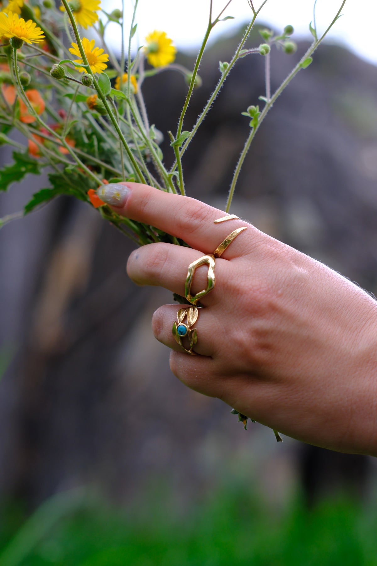 Model wears Snake Spiral Ring in 14k Gold, Split Root Ring in 14k Gold, and Willow Ring on fingers while holding a bouquet of flowers with blurred background