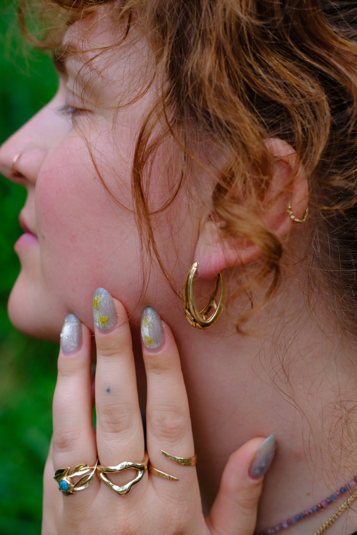 Model wears Snake Spiral Ring in 14k Gold, Split Root Ring in 14k Gold, and Willow Ring on fingers and Cicada Hoops in ears with blurred backdrop