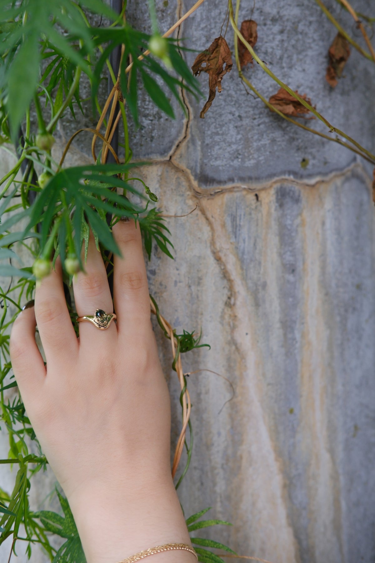 Model wears Sapphire Root Ring stacked with Root Curve Band on middle finger with blurred plants and rocks as backdrop