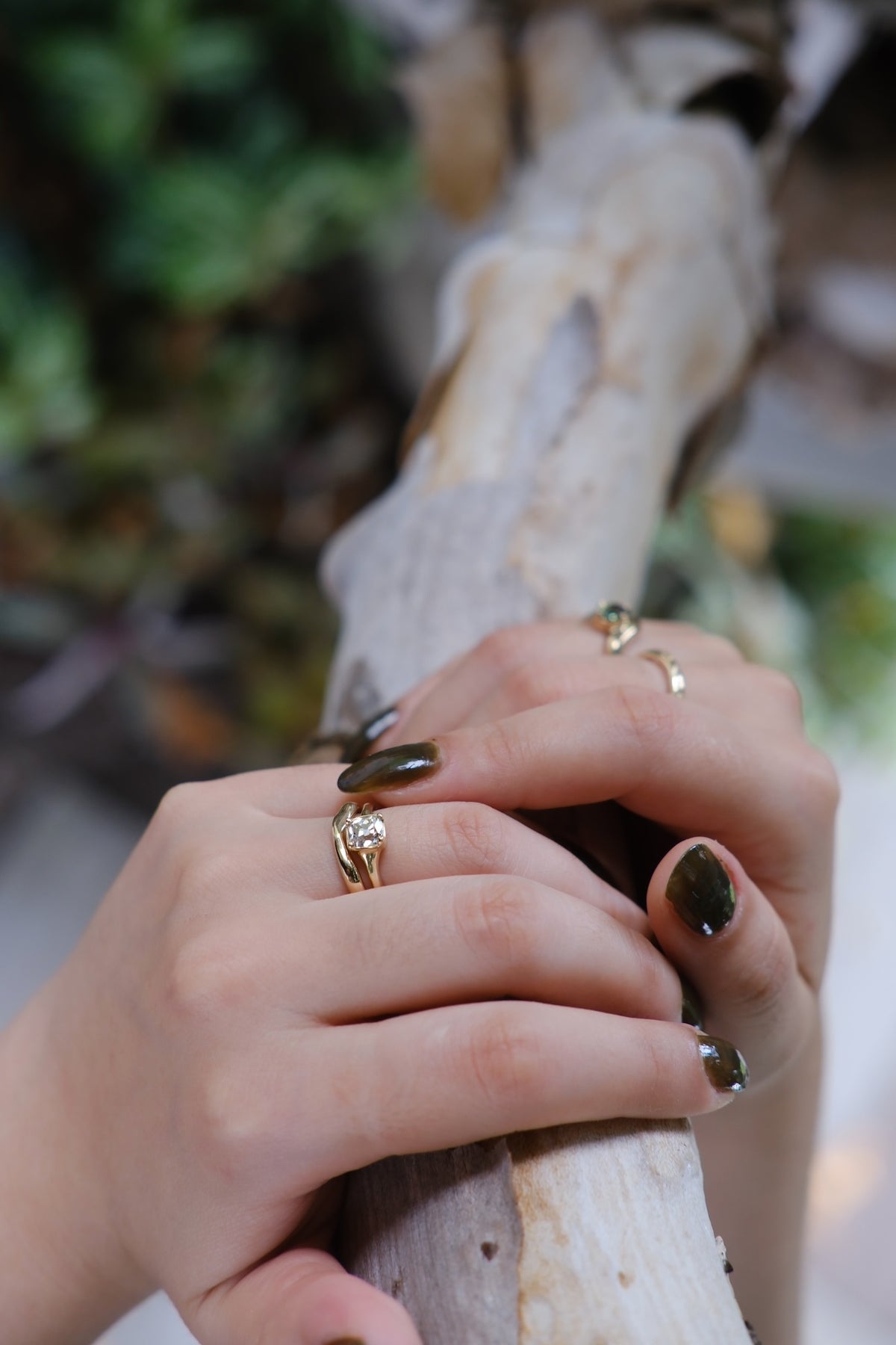 Model wears Drizzle Solitaire Ring stacked with Root Curve Band by Kestrel Dillon Jewelry on ring finger while holding a branch with blurred backdrop