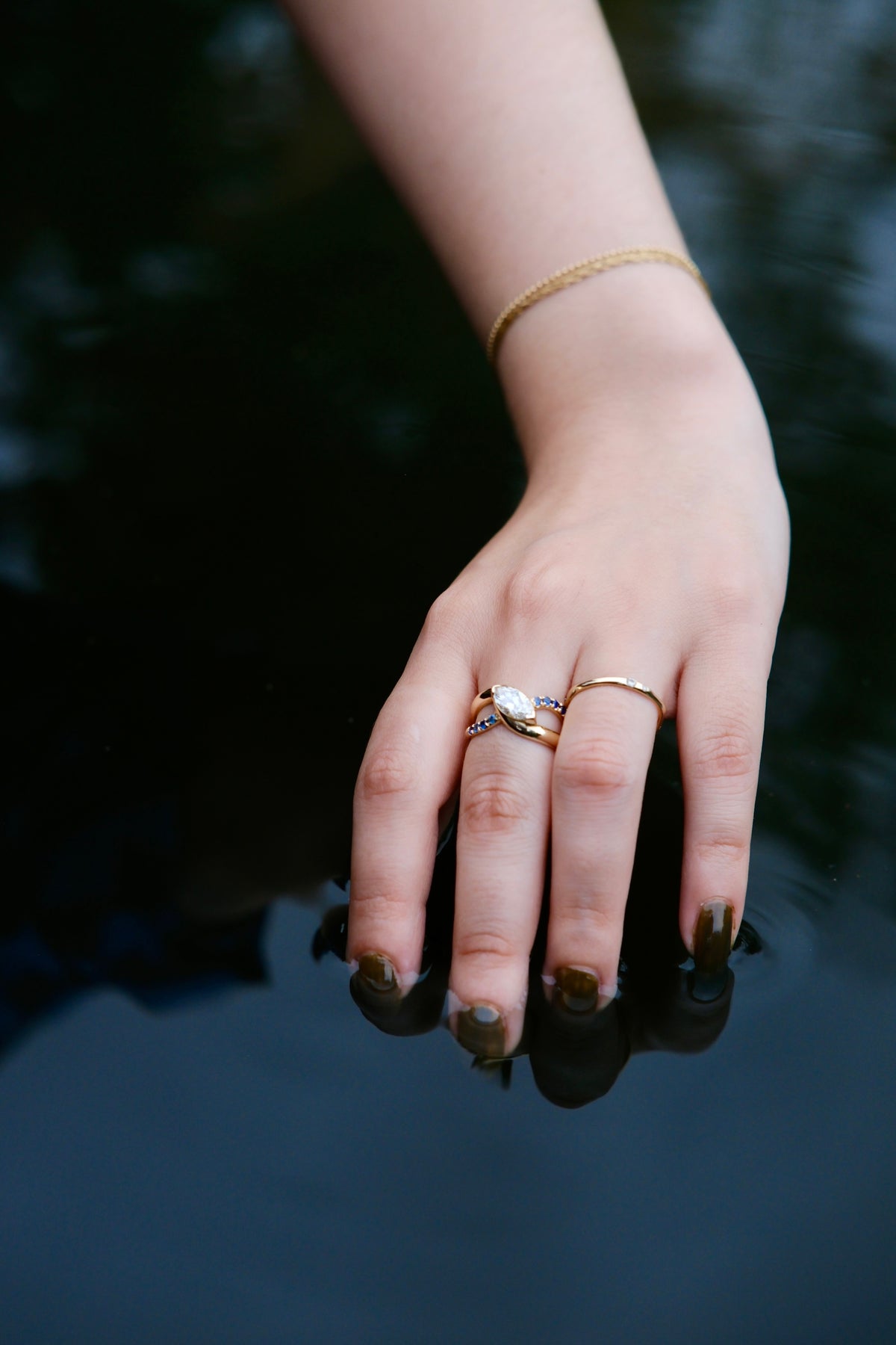 Model wears Wellspring Ring by Kestrel Dillon Jewelry on middle finger with one band on ring finger over a dark backdrop