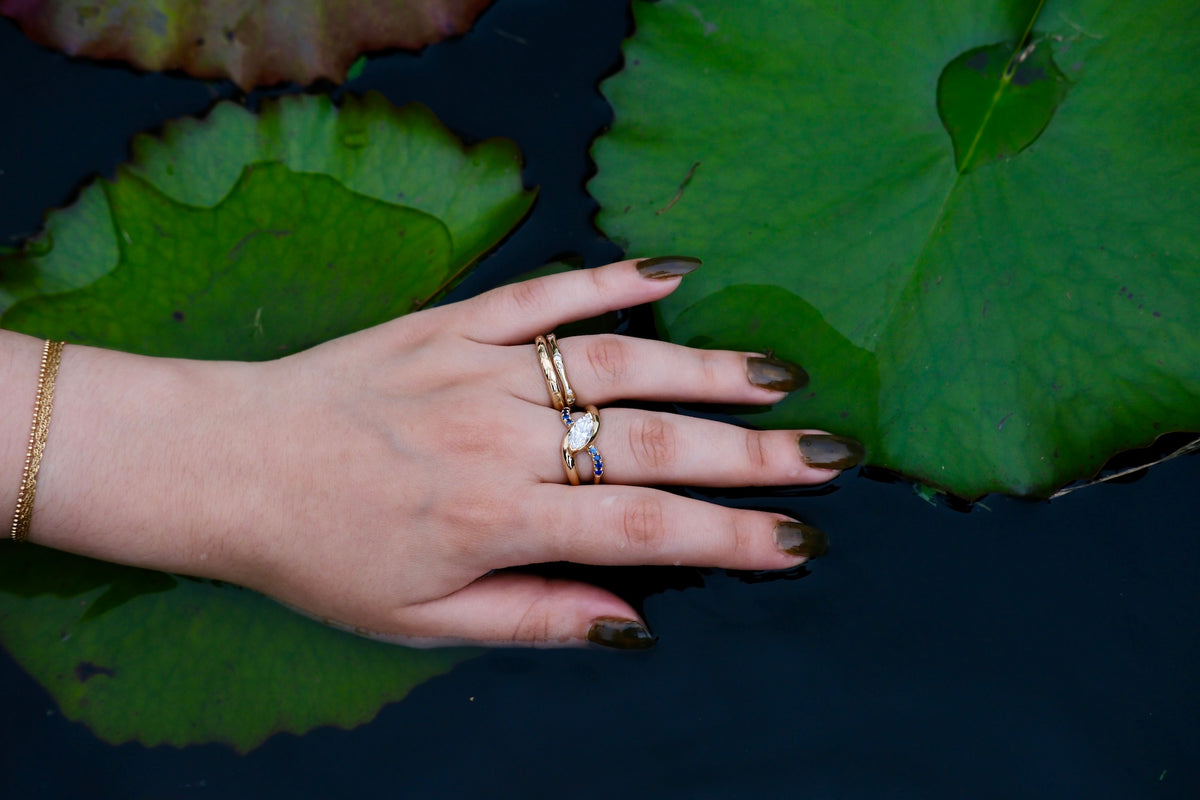 Model wears the Wellspring Ring on middle finger and two Drizzle Bands stacked on ring finger over lilly pads and dark water