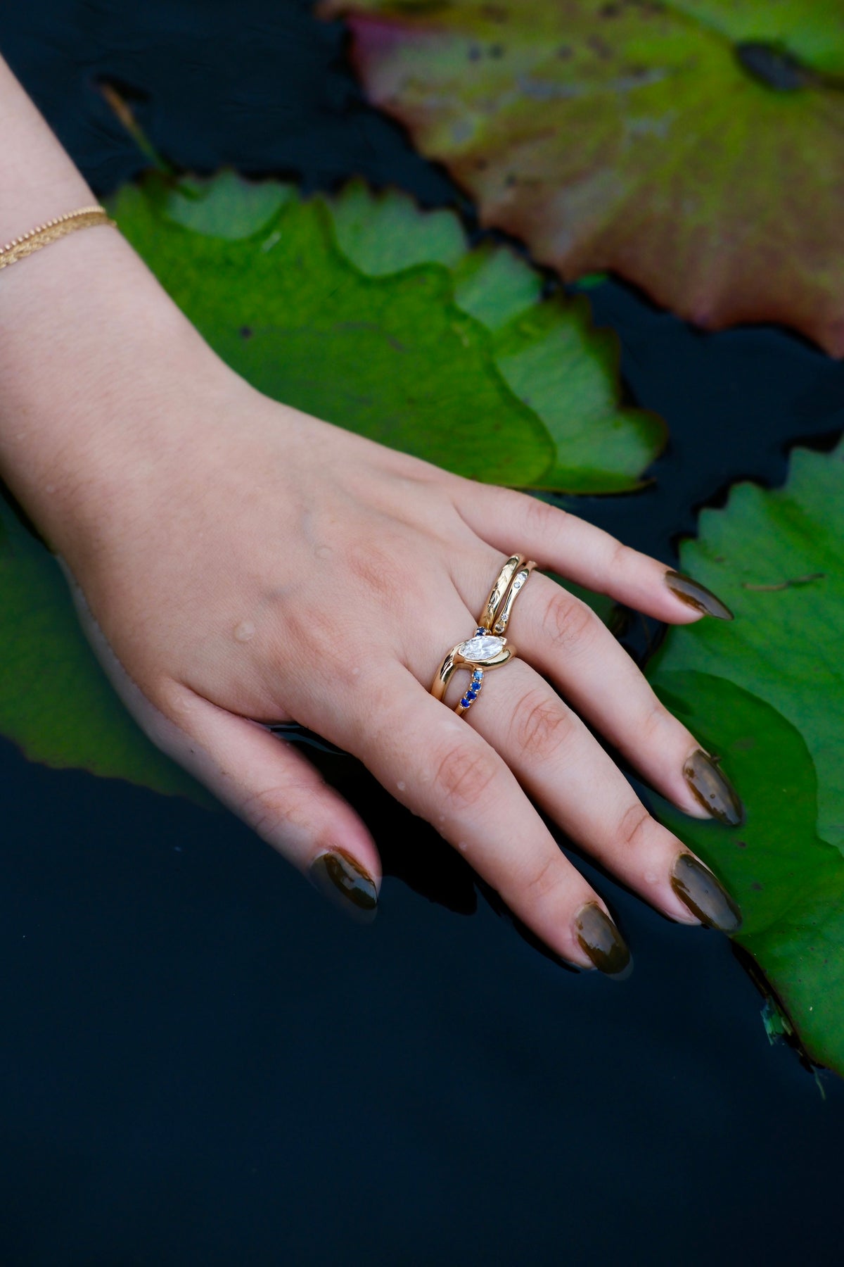 Model wears Wellspring Ring by Kestrel Dillon Jewelry on middle finger with two bands stacked on ring finger over lillly pads and a dark backdrop