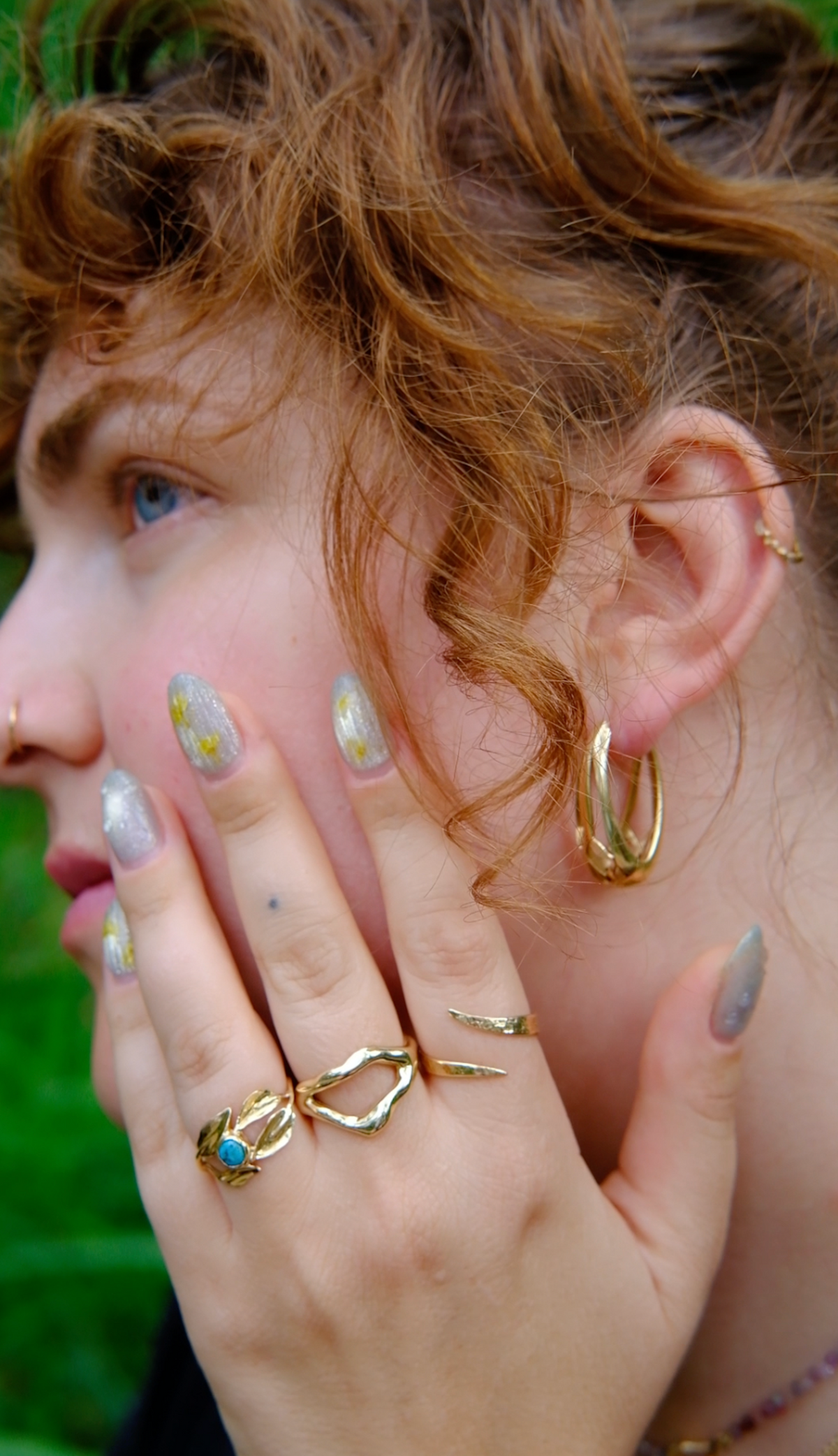 Model wears Snake Spiral Ring in 14k Gold, Split Root Ring in 14k Gold, and Willow Ring on fingers and Cicada Hoops in ears with blurred backdrop